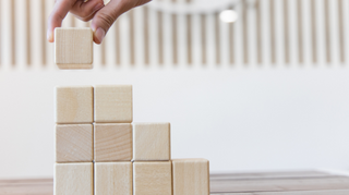 Hand stapelt auf einem Tisch kleine Holzwürfel zu einer Treppe