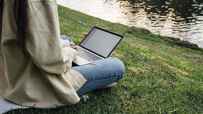 Person, die einen Laptop auf einer Wiese am Wasser benutzt.
