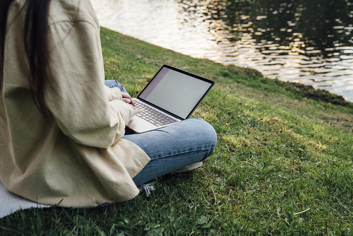Person, die einen Laptop auf einer Wiese am Wasser benutzt.