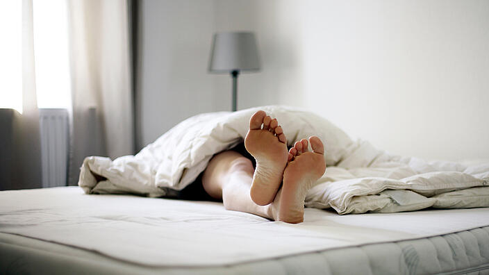 Person die auf einer Matratze liegt, die Füße schauen aus der Decke heraus