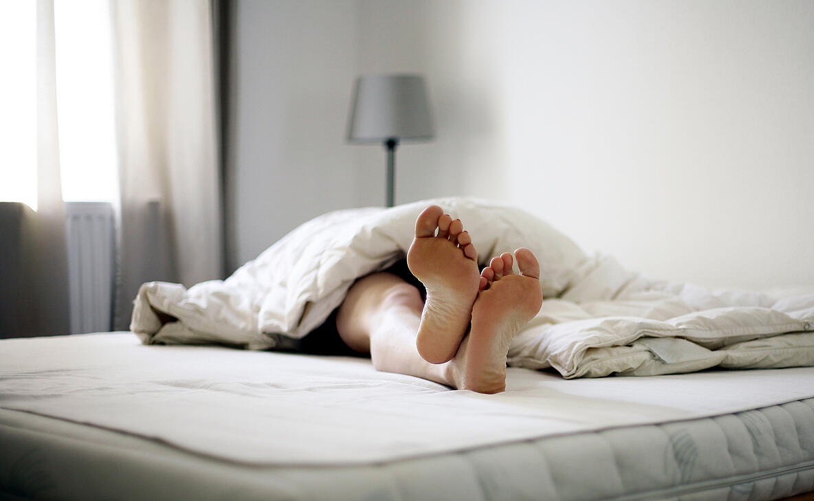 Person die auf einer Matratze liegt, die Füße schauen aus der Decke heraus