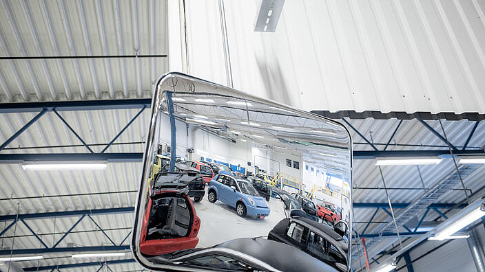 Rechteckiger Weitwinkelspiegel in einer Werkstatthalle, der mehrere Fahrzeuge und Arbeitsbereiche im Spiegelbild zeigt. Die Halle ist gut beleuchtet und hat ein Dach mit Stahlträgern.