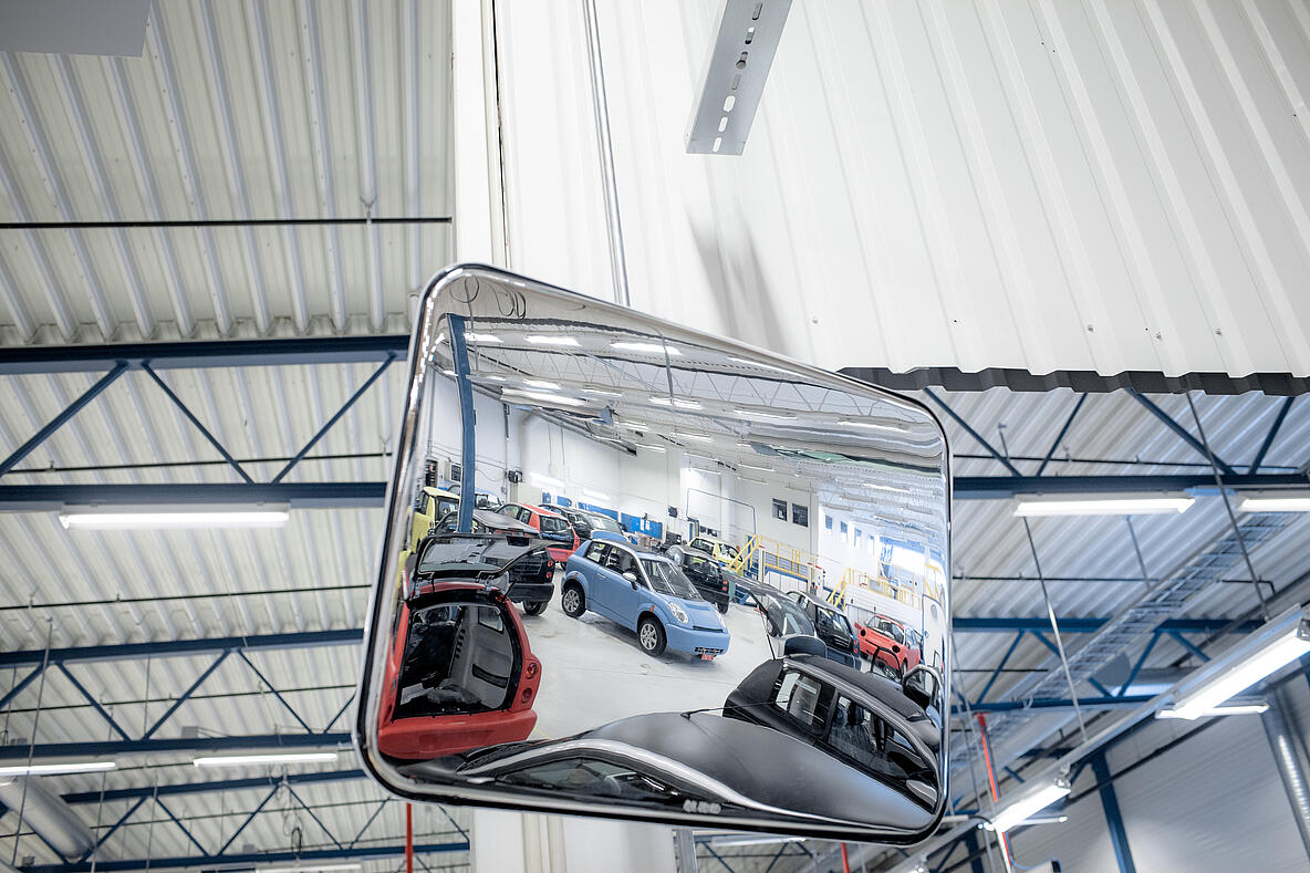 Rechteckiger Weitwinkelspiegel in einer Werkstatthalle, der mehrere Fahrzeuge und Arbeitsbereiche im Spiegelbild zeigt. Die Halle ist gut beleuchtet und hat ein Dach mit Stahlträgern.