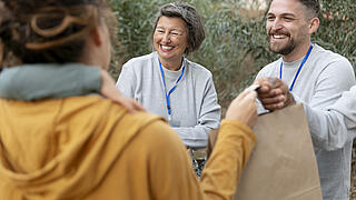 Zwei Menschen überreichen Papiertüten an eine Frau, die man von hinten sieht.