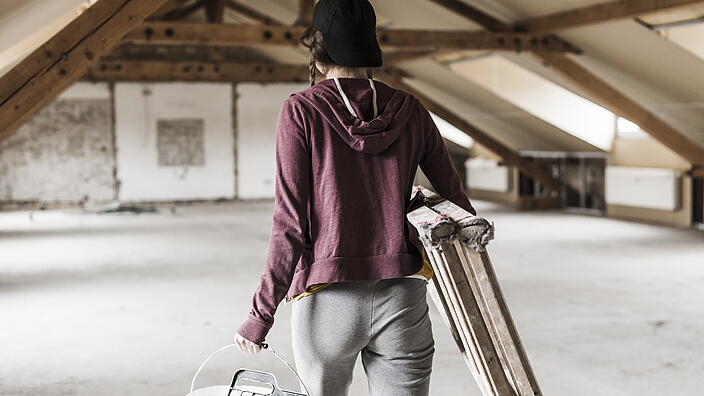 Person mit Leiter und Farbeimer auf einem Dachboden 